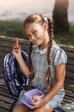 Back to school. Cute little school girl sitting on bench in school yard and eating lunch outdoor. Right school meal for lunch