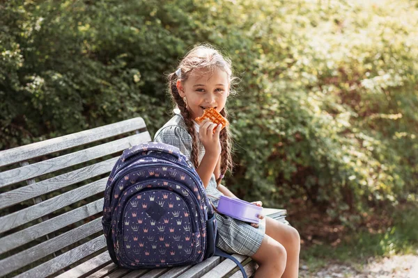 Back to school. Cute little school girl sitting on bench in school yard and eating lunch outdoor. Right school meal for lunch