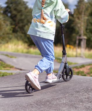  Kafkasyalı şirin bir kızın portresi güneşli bir günde asfaltlanmış Trackin Sokak Parkı 'nda scooter sürerken eğleniyor. Sağlıklı spor çocuk aktiviteleri