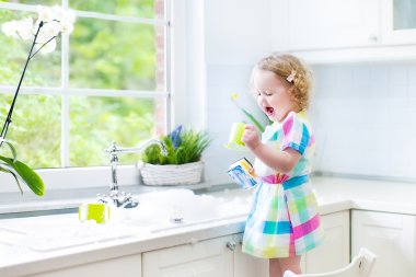 Cute curly toddler girl in a colorful dress washing dishes clipart
