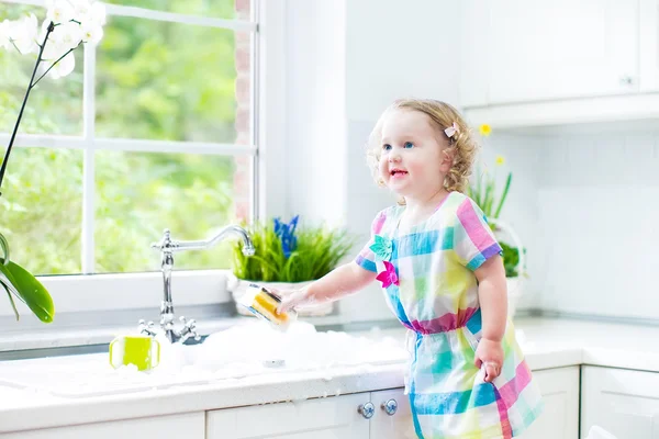 Roztomilé kudrnaté batole dívka v barevných šatech mytí nádobí — Stock fotografie