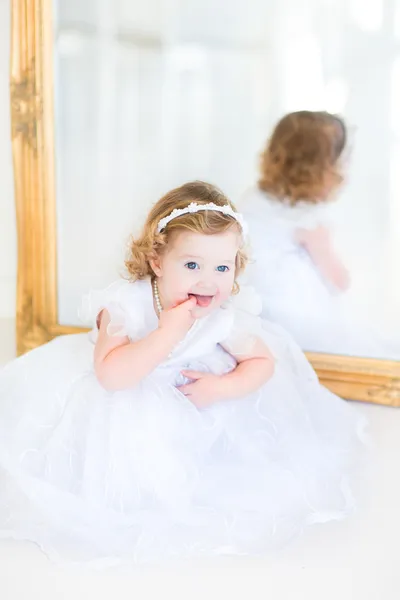 Toddler girl in a white dress sitting next to a big mirror — Stock Photo, Image