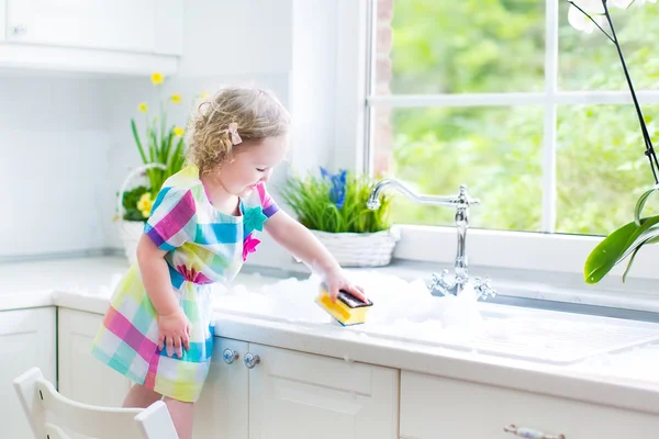 Cute curly toddler girl in a colorful dress washing dishes — Stock Photo, Image