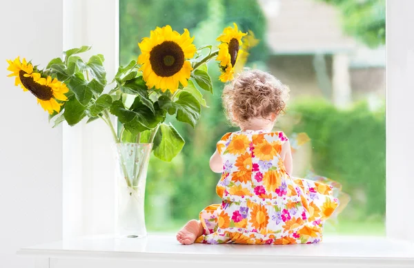 Baby girl sitting at a window next to beautiful sunflowers — Stock Photo, Image
