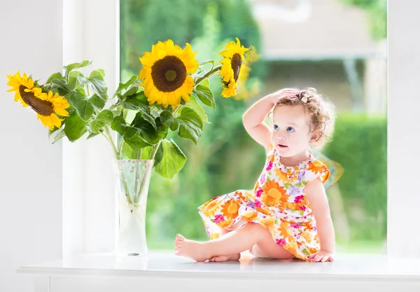 Baby girl sitting at a window next to beautiful sunflowers — Stock Photo, Image