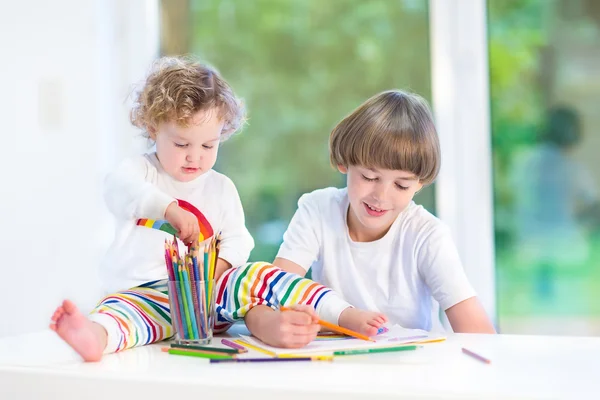 Toddler girl sitting on a white desk watching her brother drawing — Stock Photo, Image