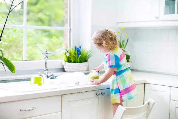 Cute curly toddler girl in a colorful dress washing dishes