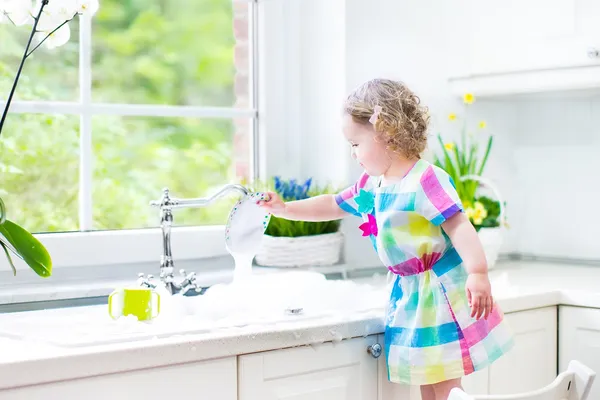 Cute curly toddler girl in a colorful dress washing dishes