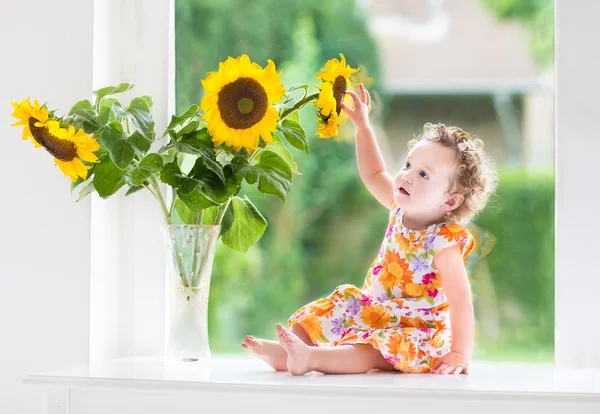 Babymeisje zit in een venster naast prachtige zonnebloemen — Stockfoto