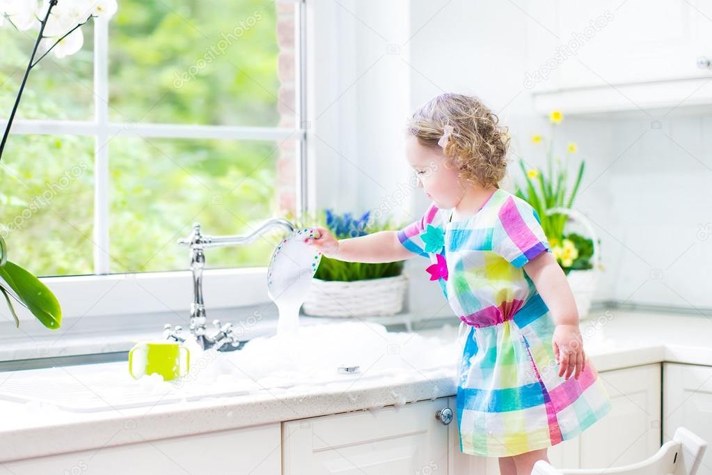 Cute curly toddler girl in a colorful dress washing dishes