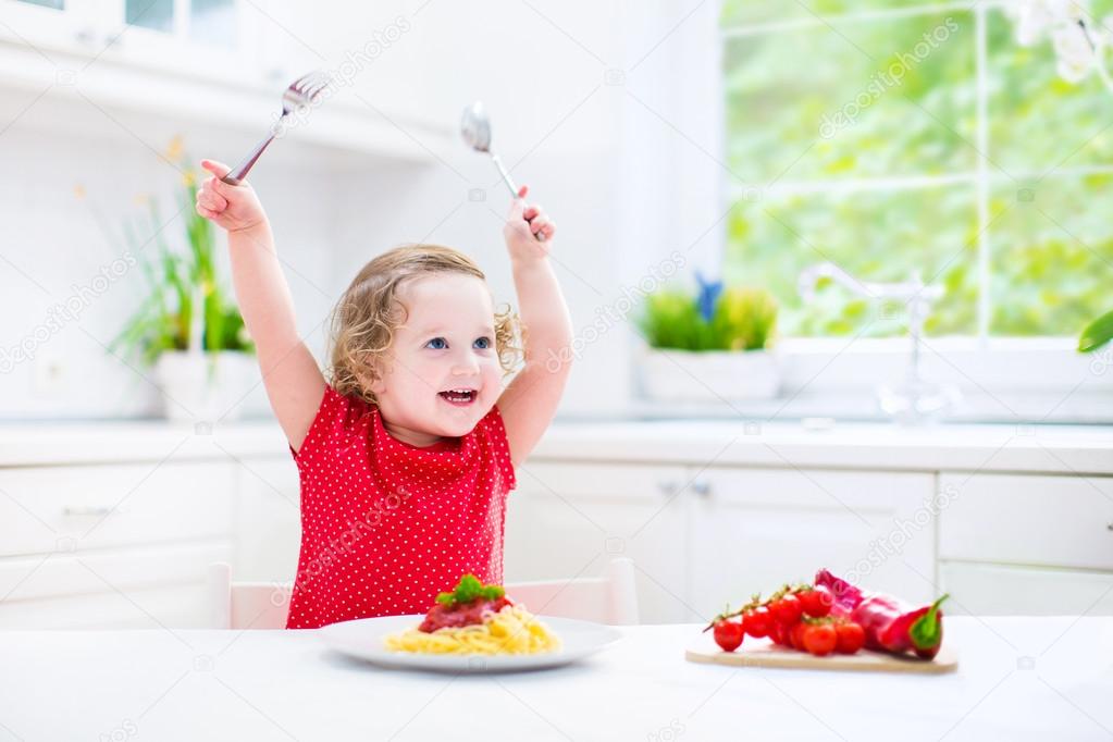 Cute toddler girl eating spaghetti in a white kitchen