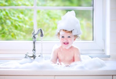 Funny baby girl playing with water and foam in a big kitchen sin clipart