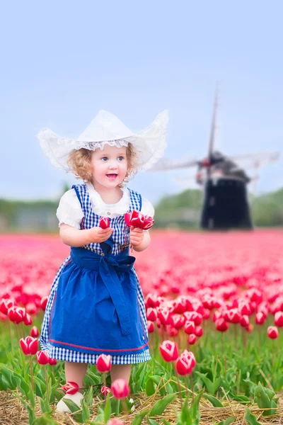 Очаровательная кудрявая девочка в голландском традиционном национальном костюме и шляпе играет в поле цветущих тюльпанов рядом с ветряной мельницей — стоковое фото