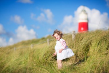 Cute toddler girl next to a red lightshouse on a beach clipart