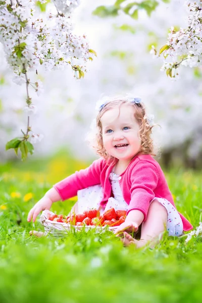 Toddler girl eating strawberry in blooming garden — Stock Photo, Image