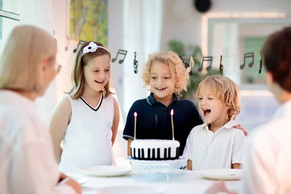 Music and piano theme kids birthday party. Kid with black and white cake. Child blowing candles and opening present. Little boy celebrating birthday. Event for young pianist. Festive decoration.