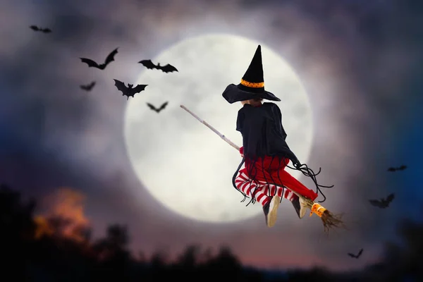 Little witch flying broom on Halloween night. Huge full moon and bats in the background. Kids trick or treat costume. Children have fun. Spooky and scary celebration. Kid dressed as evil ghost.