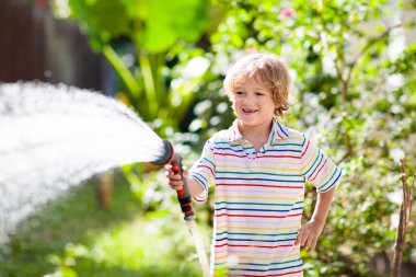 Bahçede çiçekleri ve bitkileri sulayan çocuk. Güneşli bahçesinde su hortumu olan bir çocuk. Küçük çocuk bahçeyle uğraşıyor. Evde yaz eğlencesi. Baharda aile aktivitesi. Çocuklar ebeveynlere yardım.