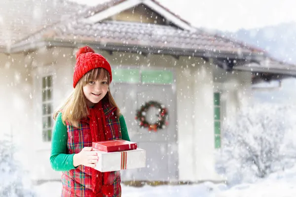 クリスマス休暇に雪の中で遊ぶ赤い帽子の子供 家族の訪問にクリスマスを提示します 冬の屋外の楽しみ 雪の公園で子供たちが遊ぶ ニットセーター スカーフ ミトンの女の子ギフトボックス — ストック写真