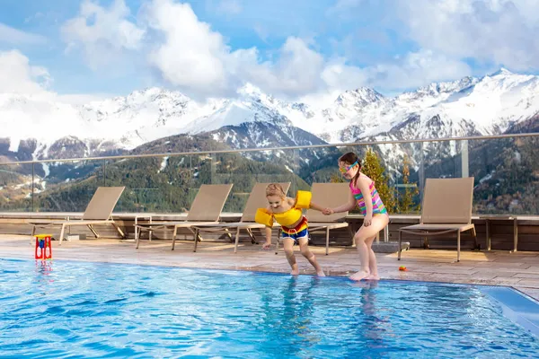 Little kids play in outdoor swimming pool of luxury alpine resort in Alps mountains, Austria. Winter and snow vacation with children. Hot tub outdoors with mountain view. Boy and girl play and swim.