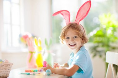 Çocuklar paskalya yumurtalarını boyuyor. Tavşan kulaklı çocuklar Paskalya avı için renkli yumurta boyuyorlar. Bahar tatili kutlaması için çiçek, sepet ve tavşanlı ev dekorasyonu. Küçük çocuk evi dekore ediyor..