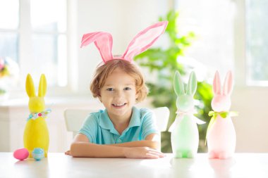 Çocuklar paskalya yumurtalarını boyuyor. Tavşan kulaklı çocuklar Paskalya avı için renkli yumurta boyuyorlar. Bahar tatili kutlaması için çiçek, sepet ve tavşanlı ev dekorasyonu. Küçük çocuk evi dekore ediyor..