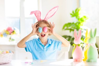 Çocuklar paskalya yumurtalarını boyuyor. Tavşan kulaklı çocuklar Paskalya avı için renkli yumurta boyuyorlar. Bahar tatili kutlaması için çiçek, sepet ve tavşanlı ev dekorasyonu. Küçük çocuk evi dekore ediyor..