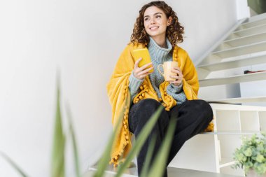 pretty curly haired woman at home in winter with a blanket and hot drink and mobile phone
