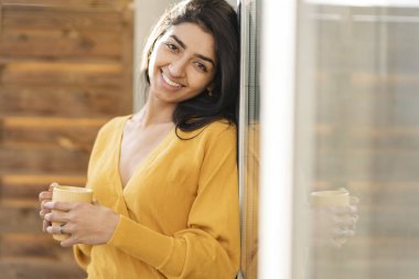 Güzel Latin kadın bir bardak içkiyle pencereye yaslanıyor.