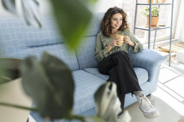 Kanepede bir fincan sıcak içecekle güzel kıvırcık saçlı kadın, ev hayatı, mutlu beyaz kadın.