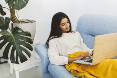 Evdeki kanepede dizüstü bilgisayarı ve battaniyesi olan İspanyol kadın.