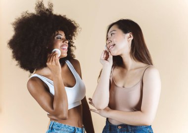 Makyaj çıkarıcı. İki güzel çok ırklı kadın, Asyalı ve Afrikalı, süngerli, izole edilmiş bej arkaplan