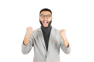 Beyaz arka planda beyaz ceketli ve gözlüklü yakışıklı Asyalı genç adam mutlu ve hareketli kazanan kollarını kaldırmış, gülümseyen ve başarı için bağıran. Kutlama