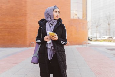 Tesettürlü genç bir kadın yoga minderiyle caddede duruyor ve modern akıllı telefon kullanıyor. Spor giyimli Müslüman bir kadın dışarıda antrenman yapıyor..