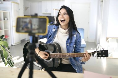Podcast sırasında gitar çalan etkili genç bir kadın veya evdeki cep telefonundan izleyiciler için canlı video yayını - konsept sanat, hobi ve video blogu