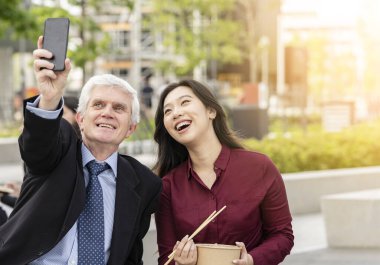 İki iş arkadaşı, kıdemli beyaz erkek ve genç Asyalı kadın cep telefonuyla gülümseyen iş adamları..