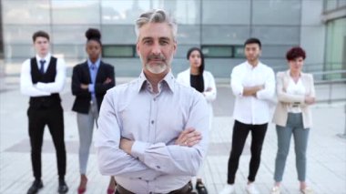 Arka planda iş arkadaşlarının açık havada yaptığı toplantıda kollarını kavuşturup gülümseyen kıdemli iş adamının videosu.