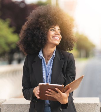 Dışarıdaki neşeli genç iş kadını tablet kullanıyor. Güzel, kıvırcık saçlı, Afro-Amerikalı bir kadın..
