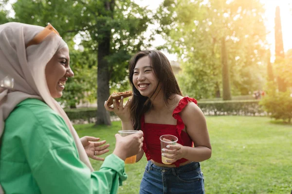 multiracial female friends eat pizza and smile in the park, focus on asian woman.