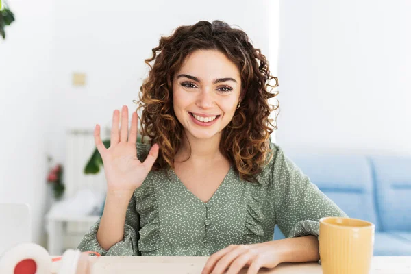 Pretty young Hispanic, vlogger woman with curly hair waving hand looking at webcam talking to camera sits at table, video call, recording blog, headshot portrait.