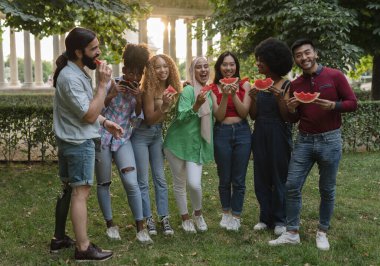 Karpuz parçalarını tutan neşeli çok ırklı dostların yakın bir portresi. Gün batımında parkta kameraya gülümsüyorlar.