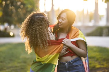 two multiethnic girlfriends with lgbt rainbow flag hugging at sunset