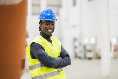 African man logistics worker in a company, with helmet and reflective vest, arms crossed smiling