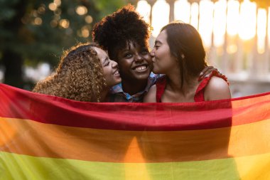 group of multiracial female friends with lgbt rainbow flag hugging at sunset