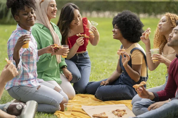 group of multiracial friends picnicking eating pizza and drinking drink, having fun in city park at sunset.