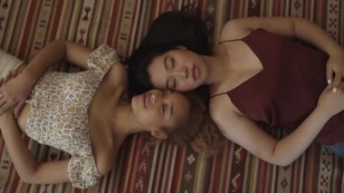 top view of two pretty multiracial women lying on the floor at home, Friendship. Two young beautiful girls on light floor looking at camera and smiling wearing pajamas