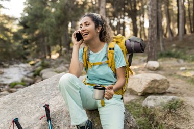 Dağlardaki sırt çantalı kadın bir fincan kahveyle cep telefonuyla gülümsüyor..