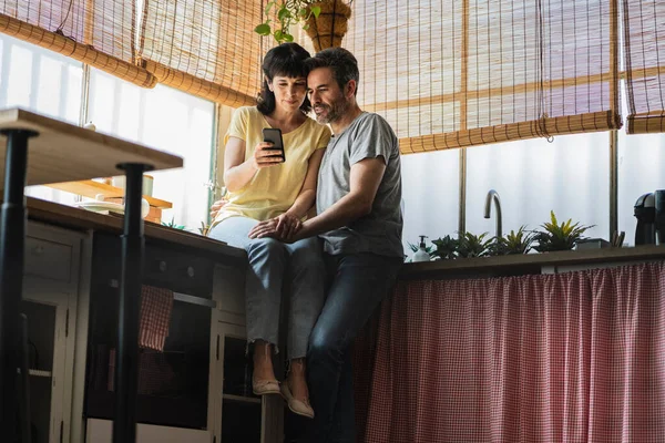 Mature woman surfing the net and enjoying with her husband using smartphone, couple sitting on kitchen counter, senior couple.