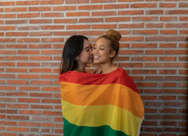 two lesbian women kiss with a gay pride rainbow flag, on a brick wall