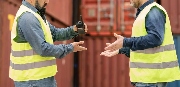 Male workers and teams in safety uniforms and helmets use walkie-talkies, work on the logistics crane with stacks of containers, control the loading of shipping goods and the freight forwarding industry.